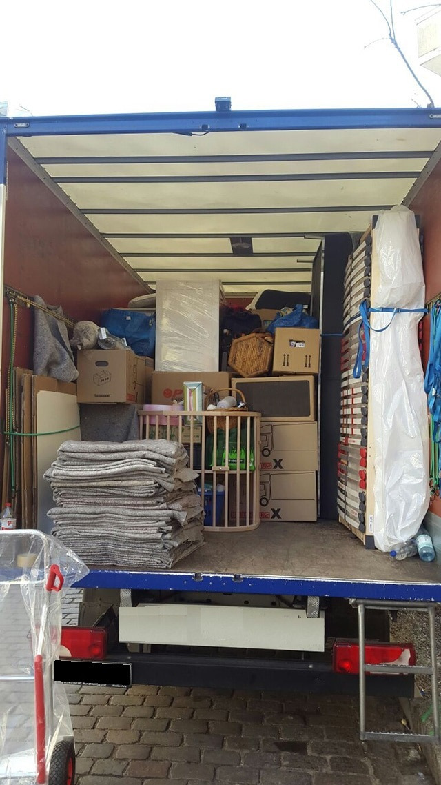 Wnętrze furgonetki wypełnionej kartonami, meblami i zabezpieczeniami, gotowej do transportu. Widoczny wózek transportowy z folią ochronną.