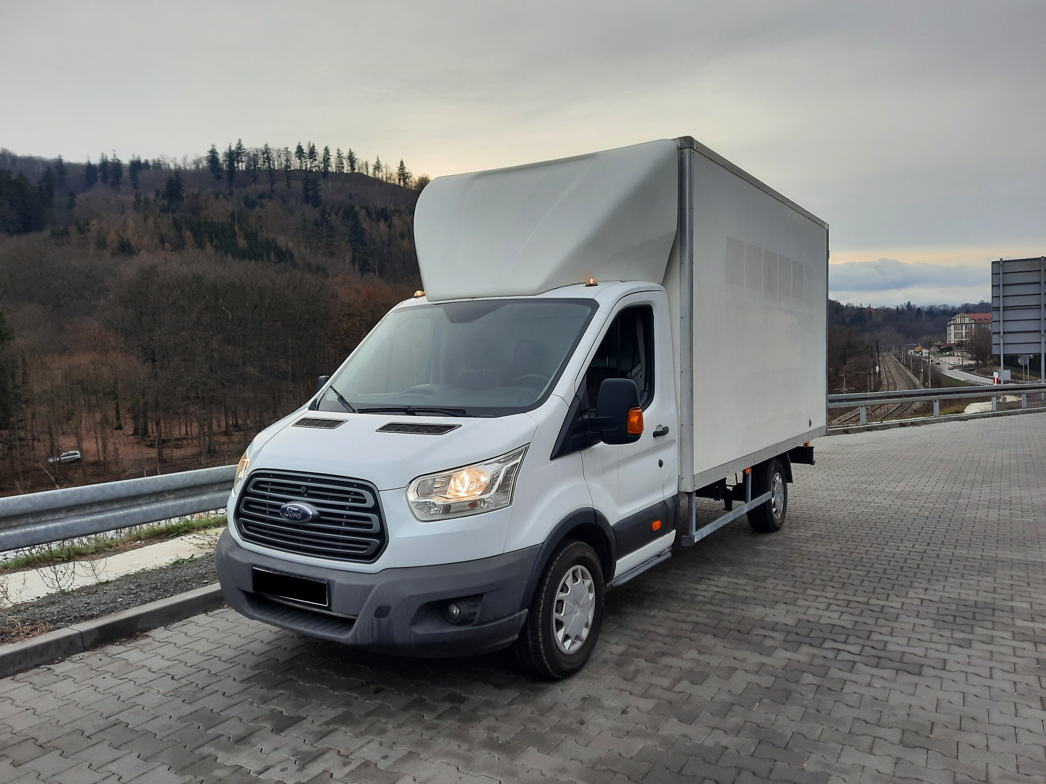 Biały bus dostawczy Ford Transit z zabudową kontenerową na tle zalesionego wzgórza i szarego nieba. Widok z boku, oświetlenie dzienne.