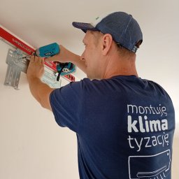 IK Klimat Ivan Ivanieiev - Mężczyzna w koszulce z napisem 'montuję klimatyzację' montuje uchwyt klimatyzatora na ścianie za pomocą wkrętarki. Widoczna poziomica i metalowy wspornik.