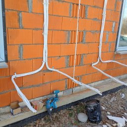 IK Klimat Ivan Ivanieiev - Montaż instalacji chłodniczej na ceglanej ścianie budynku. Białe rury przymocowane klamrami, w tle okno. Widoczne narzędzia pracy: wiertarka i młotowiertarka.