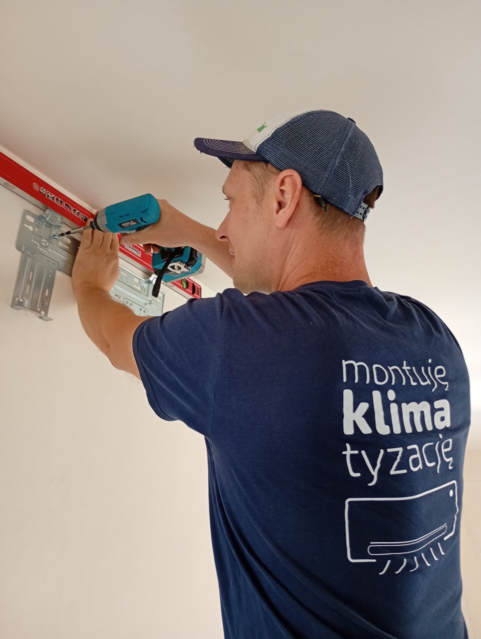 Mężczyzna w koszulce z napisem 'montuję klimatyzację' montuje uchwyt klimatyzatora na ścianie za pomocą wkrętarki. Widoczna poziomica i metalowy wspornik.