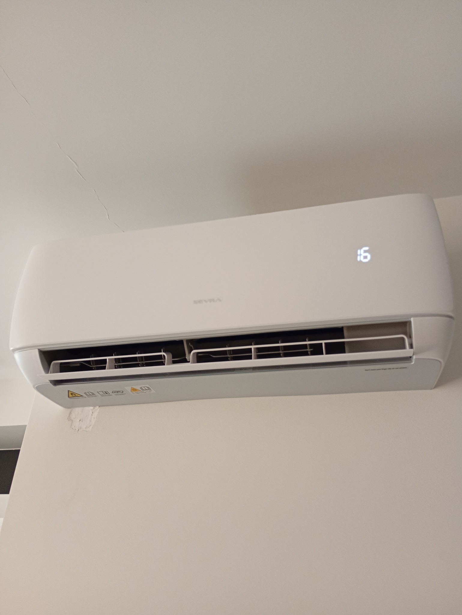 Biała klimatyzacja ścienna marki Sevra, z wyświetlaczem temperatury 16 stopni, zamontowana na jasnej ścianie. Widoczne ślady po poprzednim montażu.