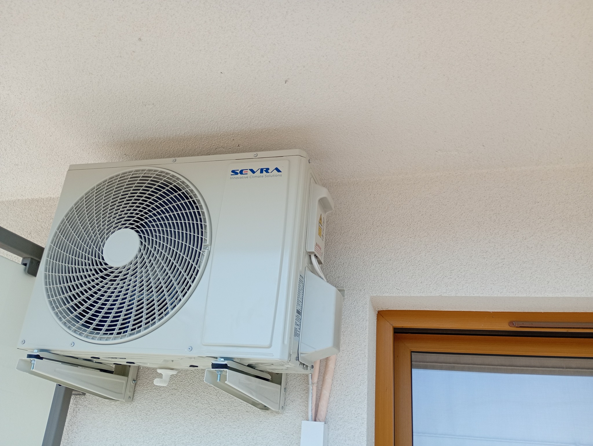 Biały klimatyzator marki Sevra zamontowany na ścianie budynku, widoczne rury i okno z drewnianą ramą. Urządzenie gotowe do użytku.