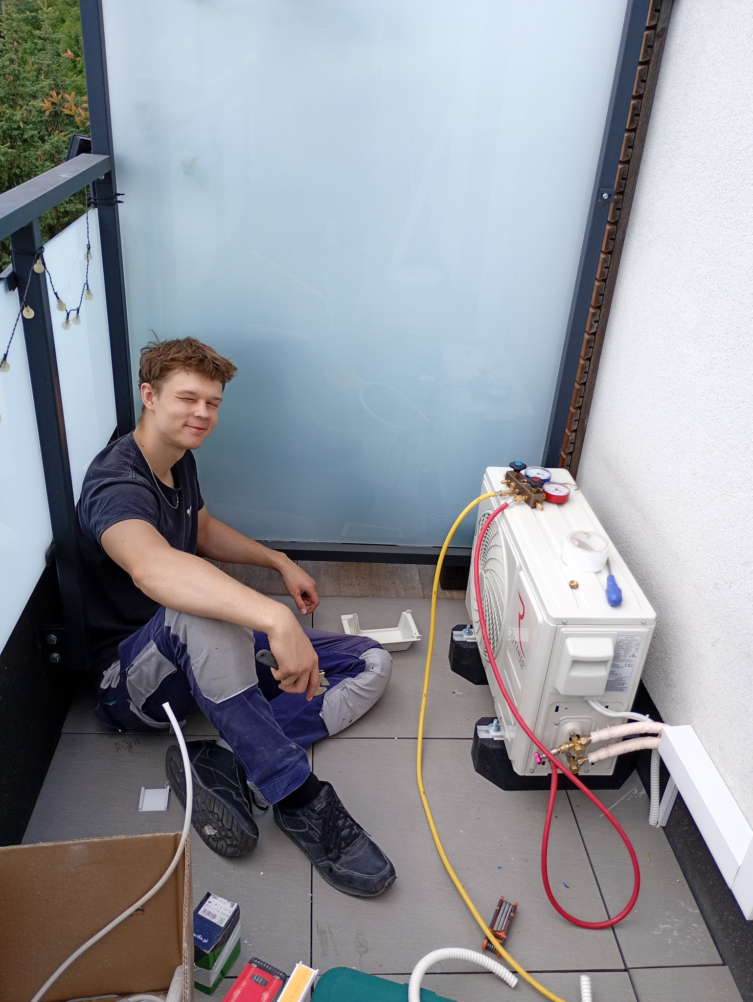 Młody monter klimatyzacji uśmiecha się podczas podłączania zewnętrznej jednostki klimatyzatora na balkonie, używając manometrów i narzędzi.