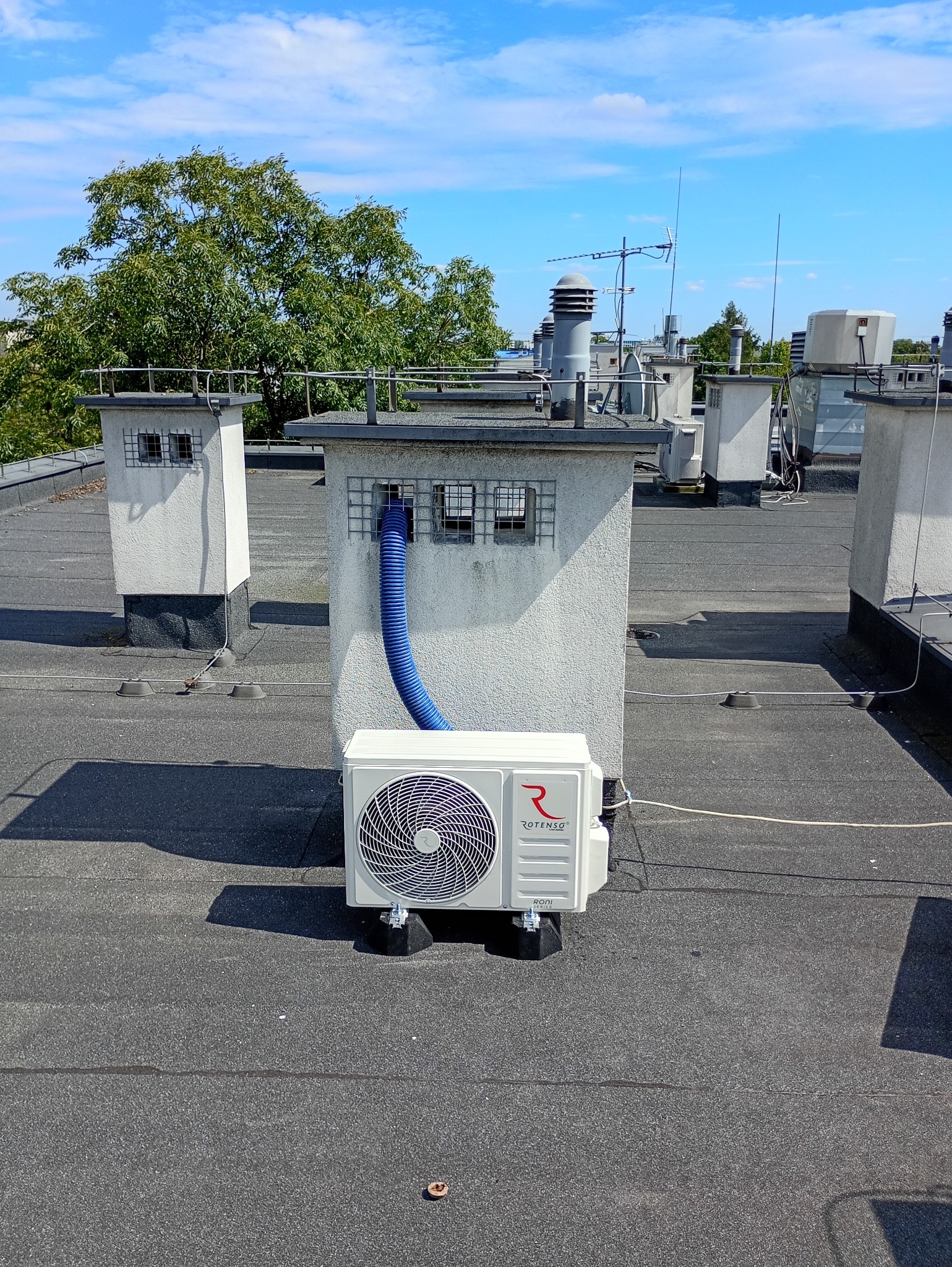 Biała jednostka zewnętrzna klimatyzacji Rotenso z niebieskim przewodem na dachu budynku, widok z góry, błękitne niebo w tle.