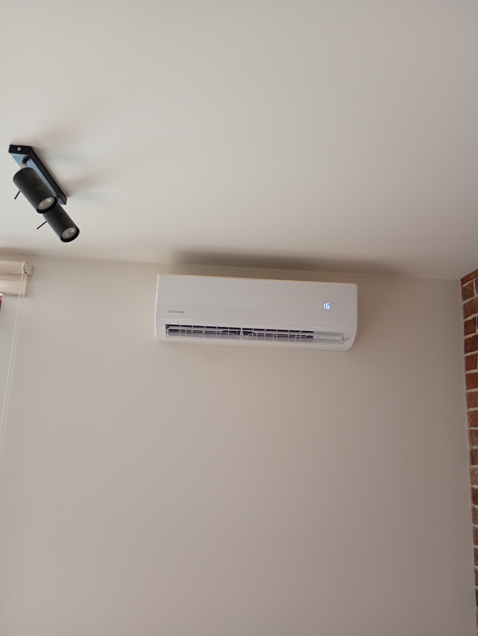 Biały klimatyzator Rotenso z wyświetlaczem temperatury (16°C) zamontowany na beżowej ścianie w pomieszczeniu z ceglanym narożnikiem i czarnym oświetleniem punktowym.