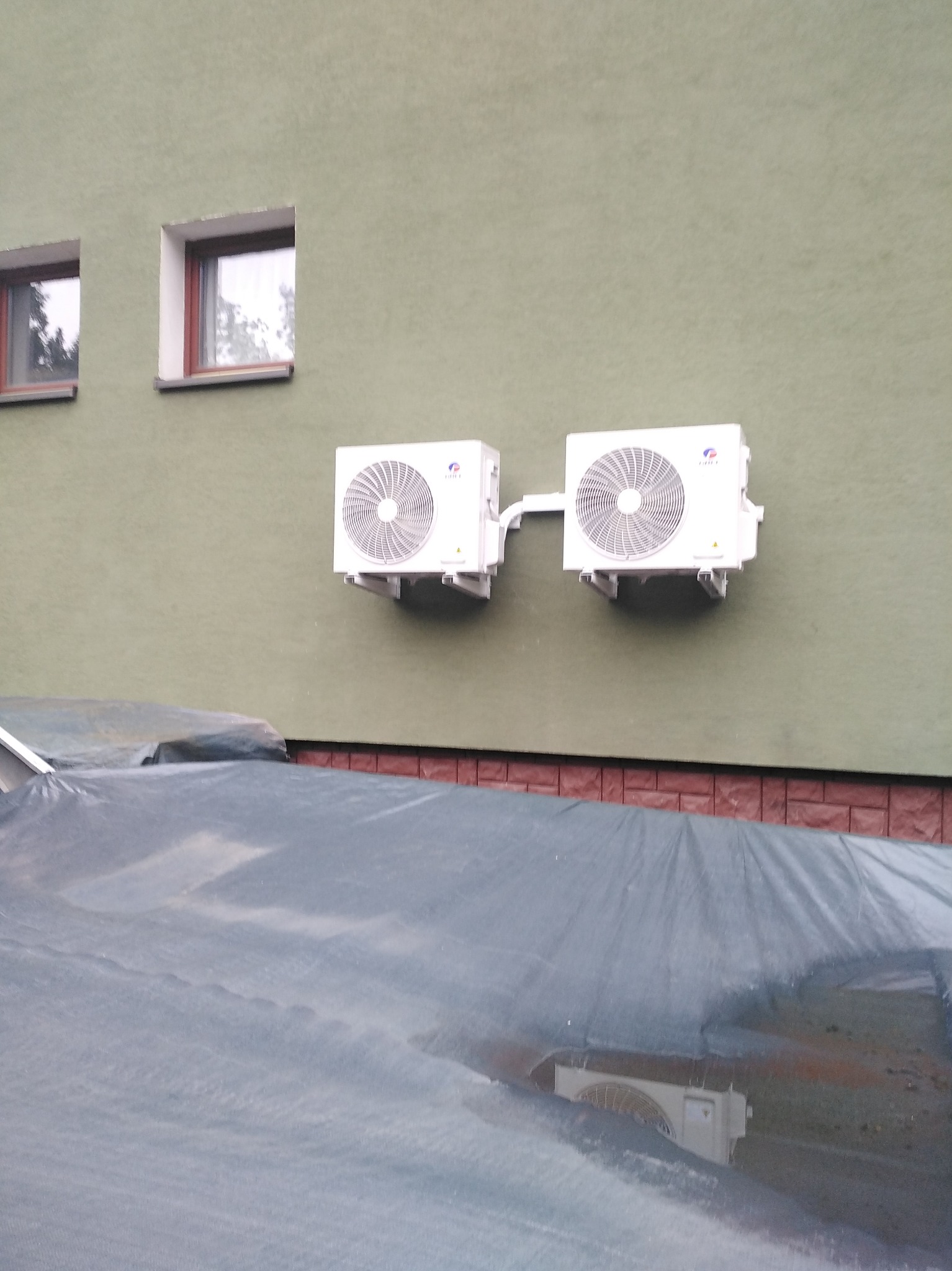 Dwa białe klimatyzatory zamontowane na zielonej ścianie budynku, widoczne spod plandeki. Widoczne dwa okna na górze. Ujęcie z dołu.