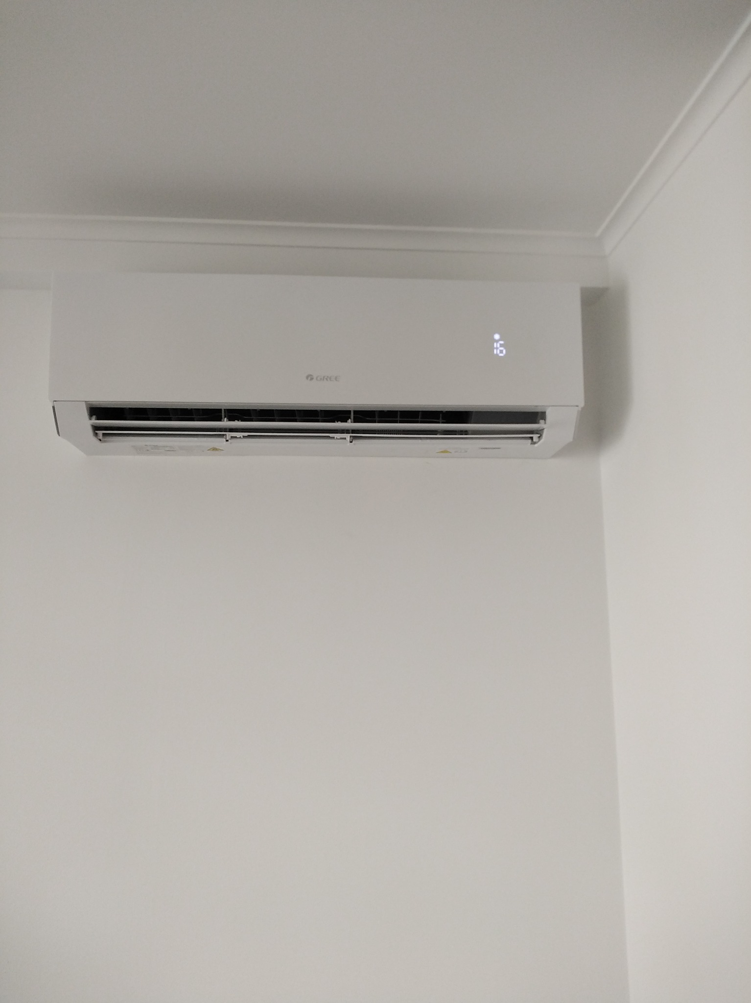 Biały klimatyzator marki Gree zamontowany na jasnej ścianie w rogu pomieszczenia, widoczny wyświetlacz z temperaturą 16 stopni.