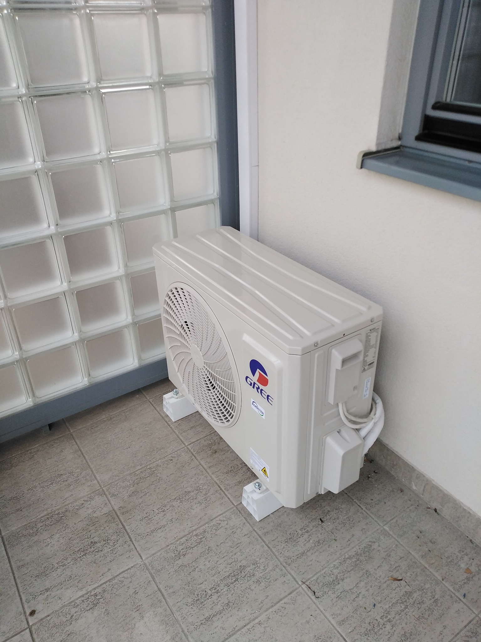 Zewnętrzna jednostka klimatyzacji Gree, zamontowana na balkonie w Piasecznie. Widoczna ściana z luksferów i fragment okna. Biała obudowa, szare płytki podłogowe.