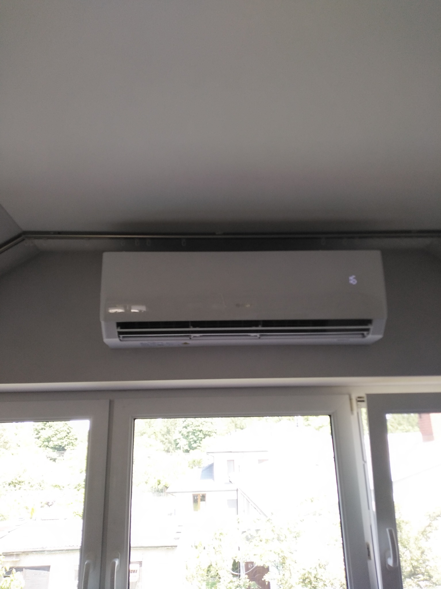 Biały klimatyzator ścienny zamontowany nad oknem, widoczny wyświetlacz temperatury. Szare ściany i sufit. Widok na zewnątrz przez okno.