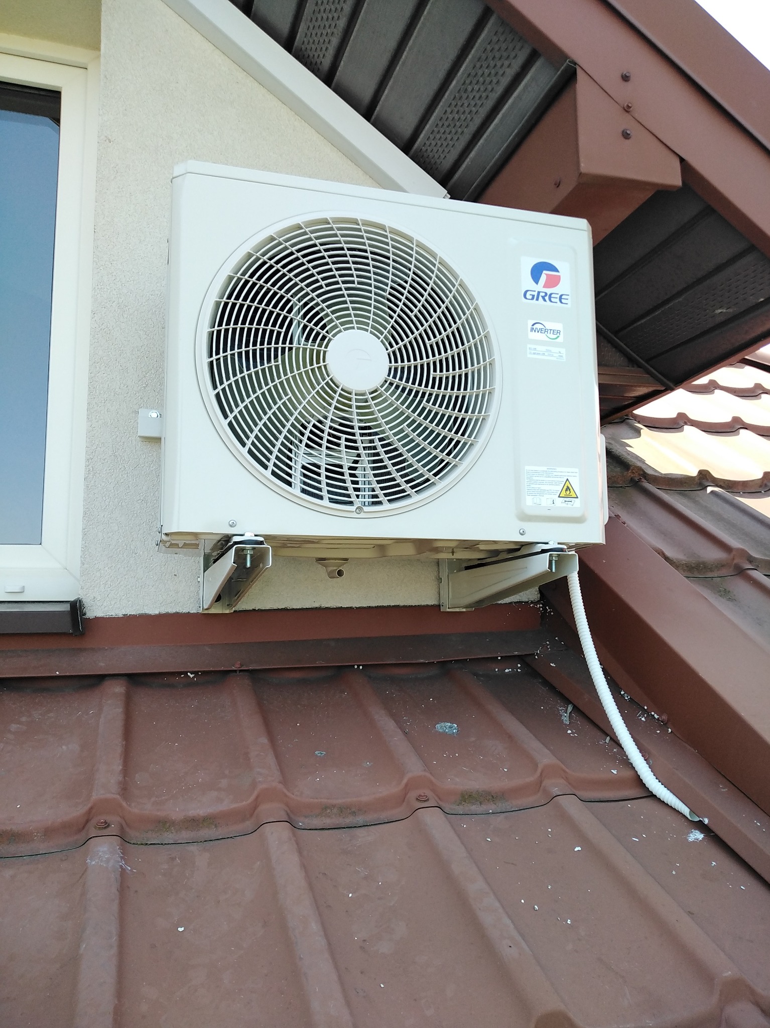 Biały klimatyzator marki Gree zamontowany na brązowym dachu z blachy, widoczny fragment okna i elewacji budynku. Kabel odprowadzający skropliny.
