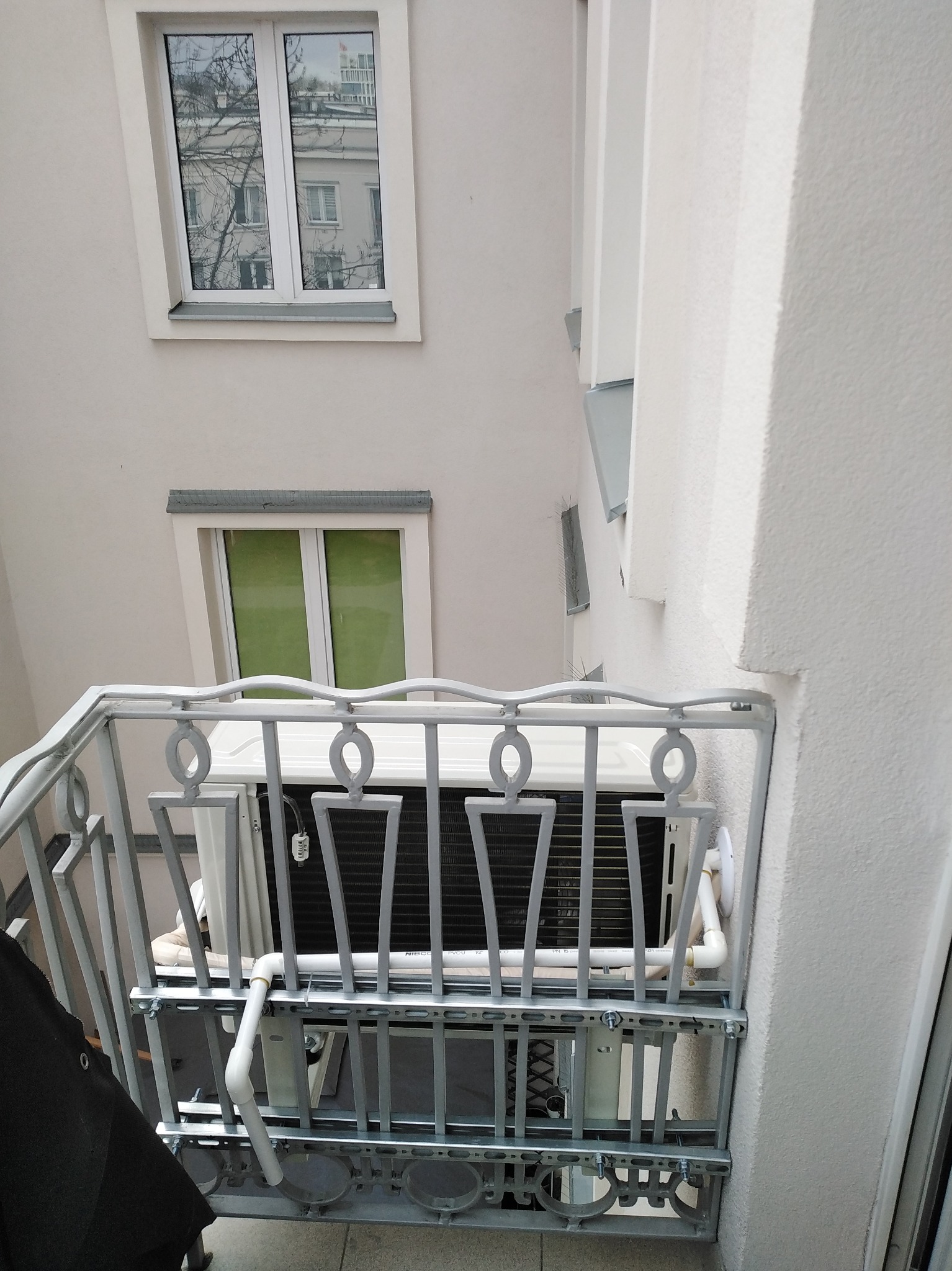 Biała jednostka zewnętrzna klimatyzacji zamontowana na balkonie z ozdobną, metalową balustradą. Widok z dołu, na tle jasnej elewacji budynku z oknami.