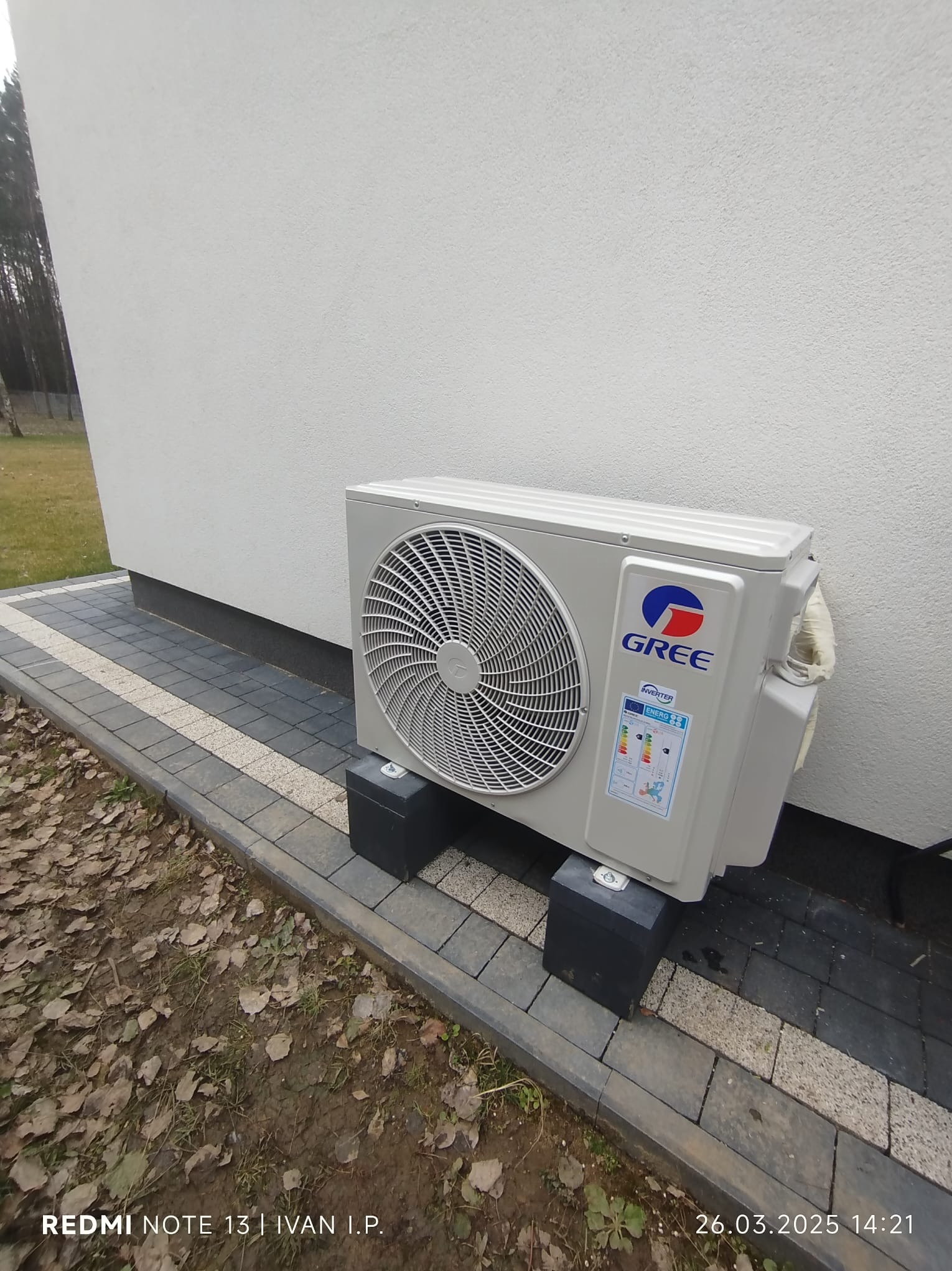 Zewnętrzna jednostka klimatyzacji Gree, zamontowana na czarnych podstawkach przy białej ścianie budynku. Widoczna kostka brukowa i fragment trawnika z liśćmi.