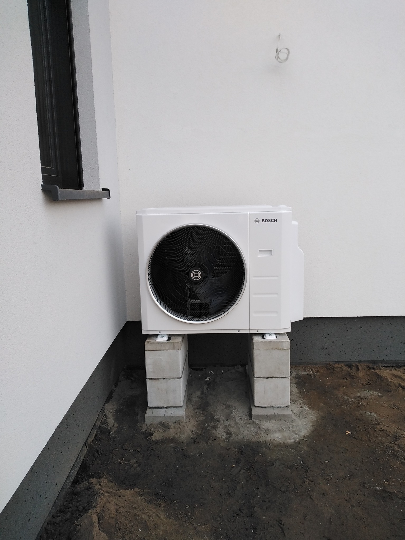 Biała jednostka zewnętrzna klimatyzacji Bosch zamontowana na betonowych bloczkach przy ścianie budynku z widocznym oknem. Świeżo wykopana ziemia wokół podstawy.