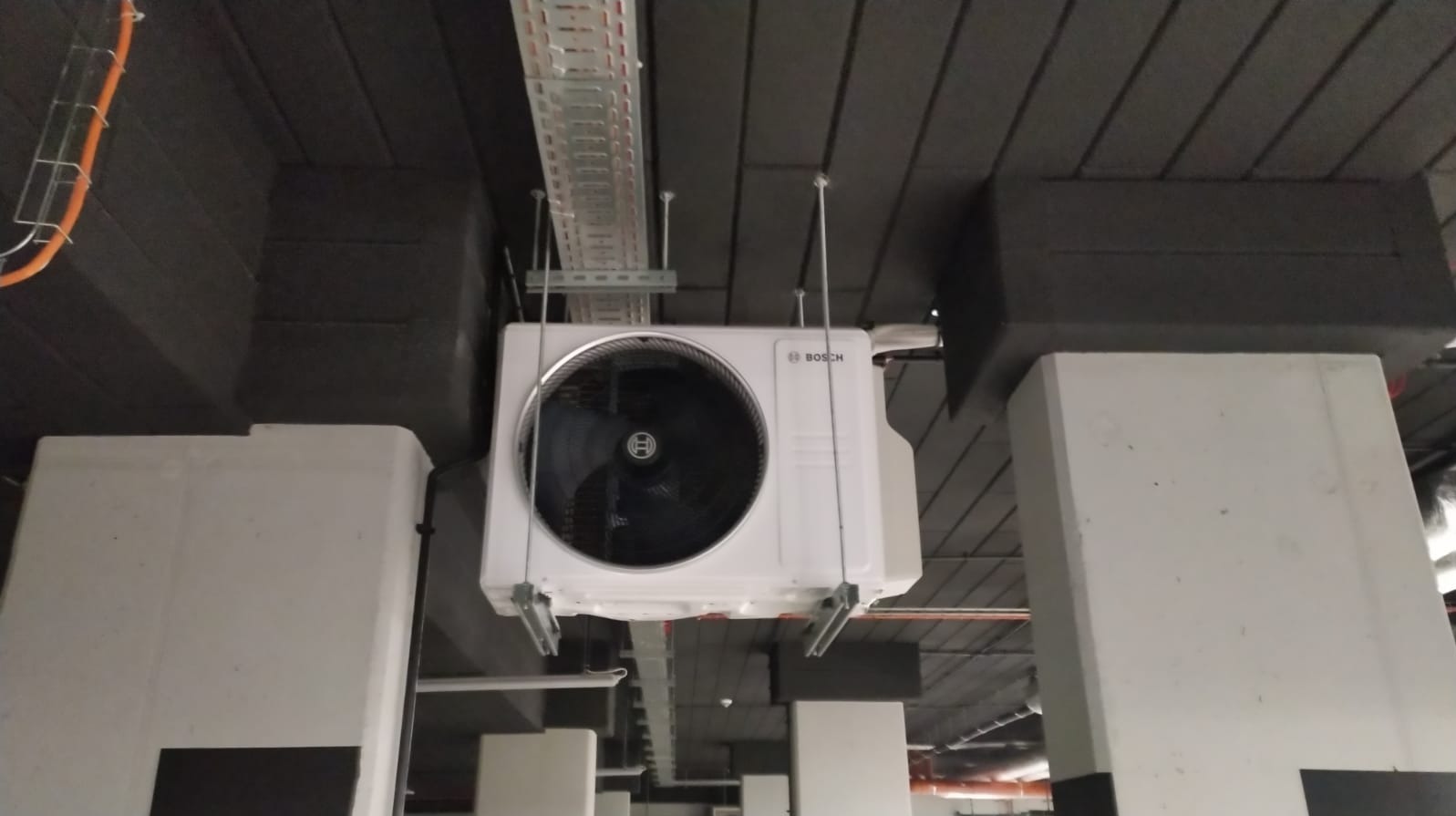 Biała jednostka klimatyzacyjna marki Bosch zamontowana pod sufitem, widoczne elementy instalacji i kanały wentylacyjne. Ujęcie z dołu, perspektywa podkreśla wysokość pomieszczenia.