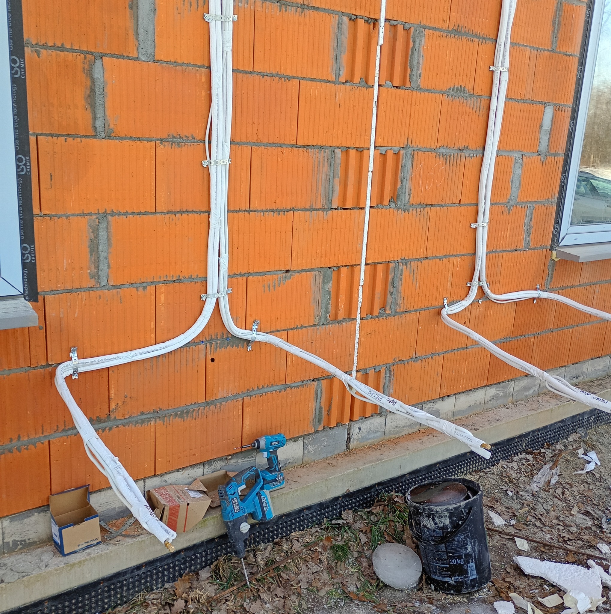 Montaż instalacji chłodniczej na ceglanej ścianie budynku. Białe rury przymocowane klamrami, w tle okno. Widoczne narzędzia pracy: wiertarka i młotowiertarka.