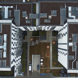 WIĘCEJ Nieruchomości - Widok z drona na nowoczesny budynek mieszkalny z dziedzińcem, zielonym dachem i charakterystyczną fasadą w czarno-białą szachownicę. Widoczne instalacje na dachu i zaparkowane samochody.