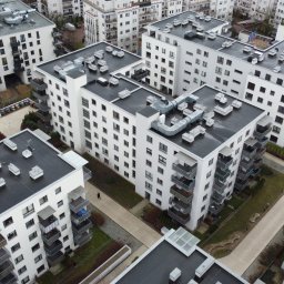 WIĘCEJ Nieruchomości - Nowoczesne, białe budynki wielorodzinne z balkonami, widok z góry. Na dachach widoczne elementy wentylacji. Pomiędzy budynkami ścieżki i zieleń.