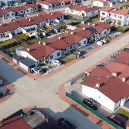 WIĘCEJ Nieruchomości - Osiedle domów jednorodzinnych z czerwonymi dachami, widok z góry. Uliczki z kostki brukowej, zadbane ogródki, samochody na podjazdach. Słoneczny dzień.