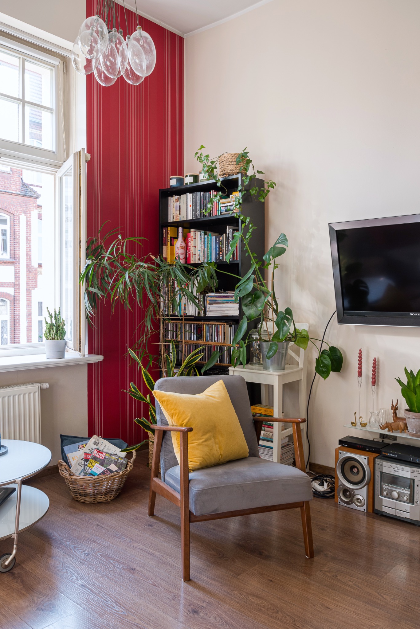 Przytulne wnętrze pokoju z szarym fotelem, żółtą poduszką, regałem z książkami i roślinami. Widoczne okno z widokiem na budynek z czerwonej cegły.