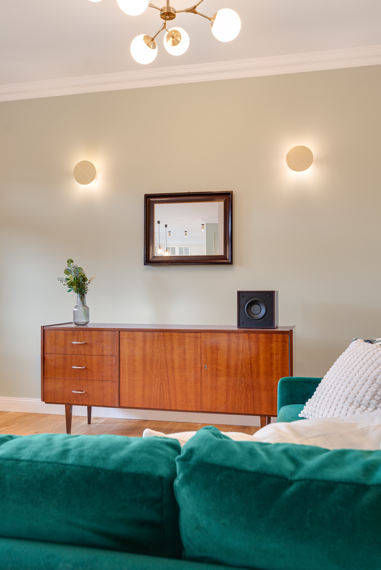 Salon z komodą w stylu vintage, lustrem w drewnianej ramie i zieloną sofą. Na komodzie głośnik i wazon z kwiatami. Oświetlenie punktowe i lampa sufitowa.