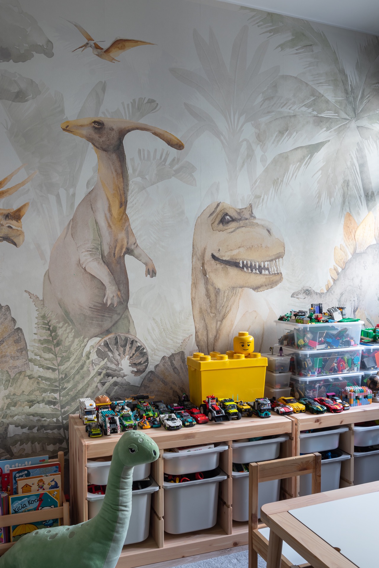 Pokój dziecięcy z tapetą w dinozaury, drewnianymi regałami wypełnionymi zabawkami, klockami Lego i pluszowym dinozaurem. Jasne, naturalne światło.