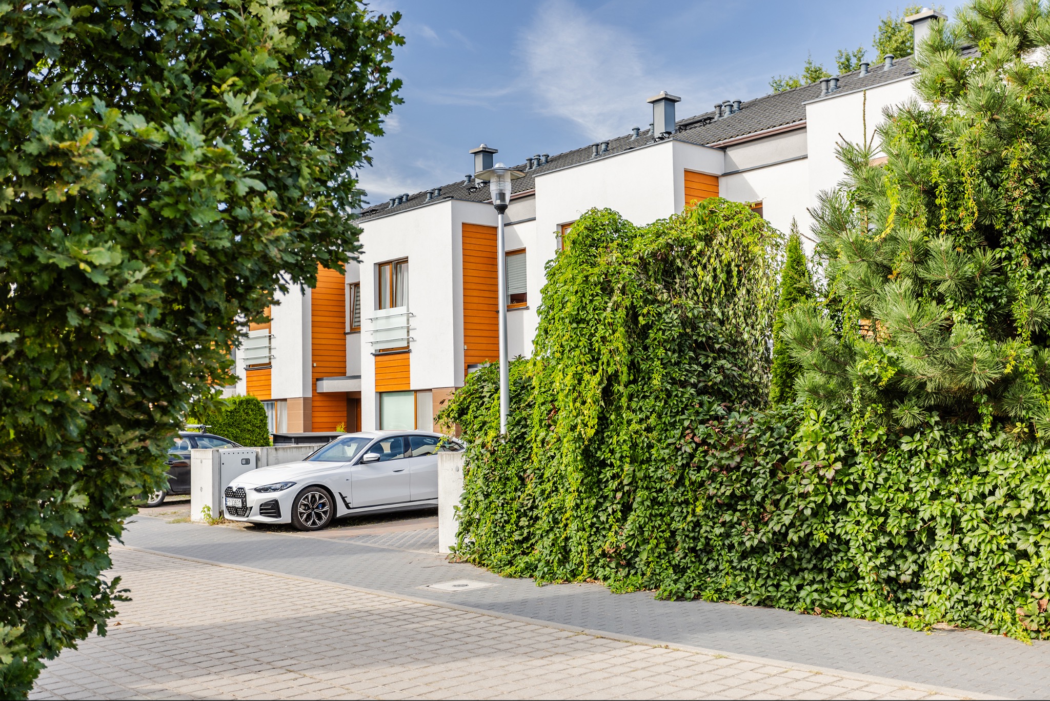 Nowoczesne osiedle z białymi domami i pomarańczowymi akcentami, bujna zieleń, zaparkowany biały samochód. Słoneczny dzień, błękitne niebo.