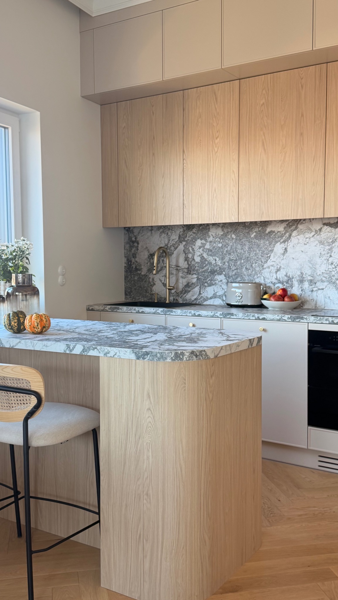 Jasna kuchnia z drewnianymi szafkami i marmurowym blatem. Na blacie dynie ozdobne, kwiaty w wazonie i owoce. Złota bateria i sprzęt AGD w stylu retro.