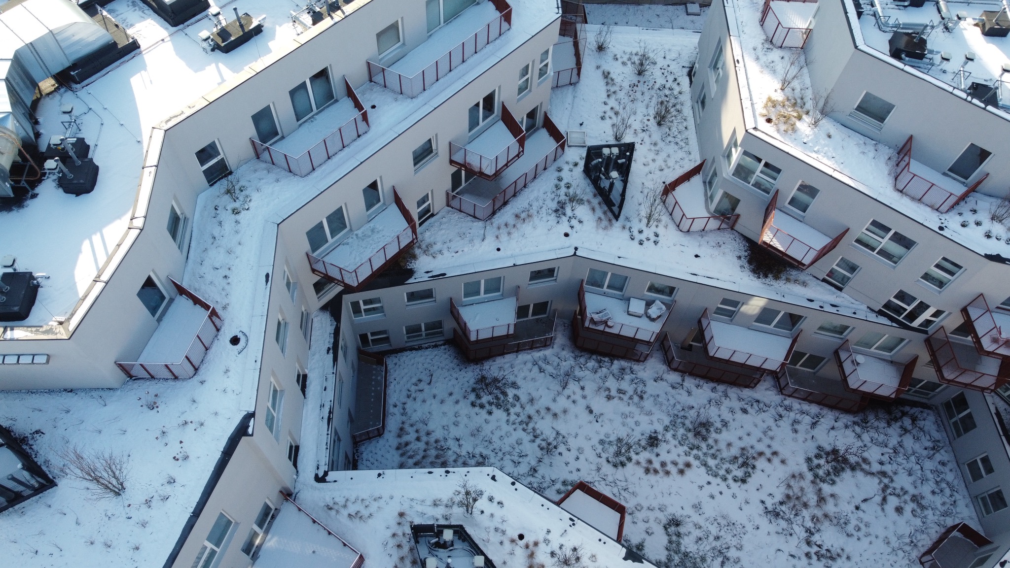 Zimowy widok z góry na osiedle z balkonami, pokryte śniegiem dachy i dziedziniec. Nietypowy kształt budynków i perspektywa z drona.