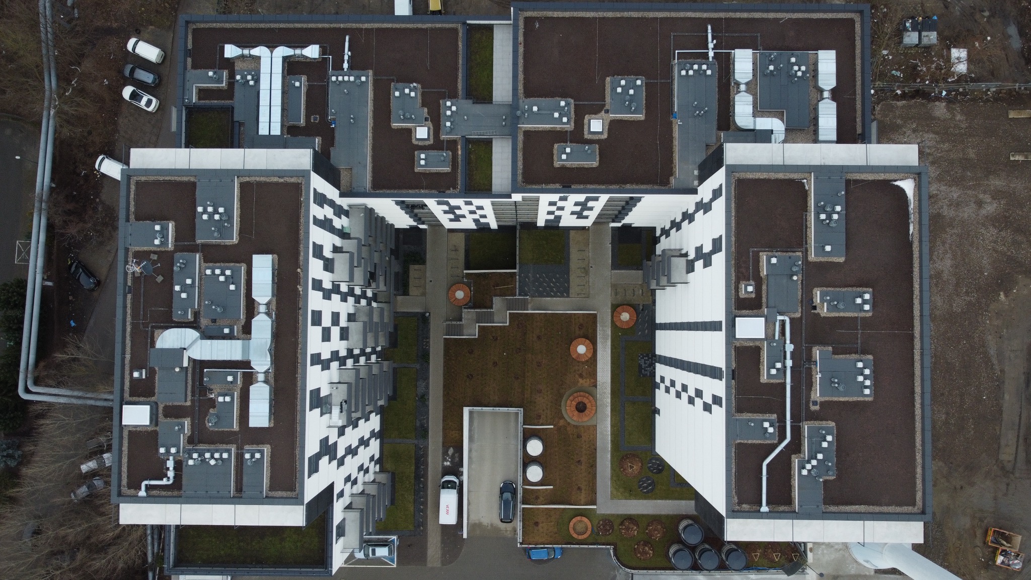 Widok z drona na nowoczesny budynek mieszkalny z dziedzińcem, zielonym dachem i charakterystyczną fasadą w czarno-białą szachownicę. Widoczne instalacje na dachu i zaparkowane samochody.