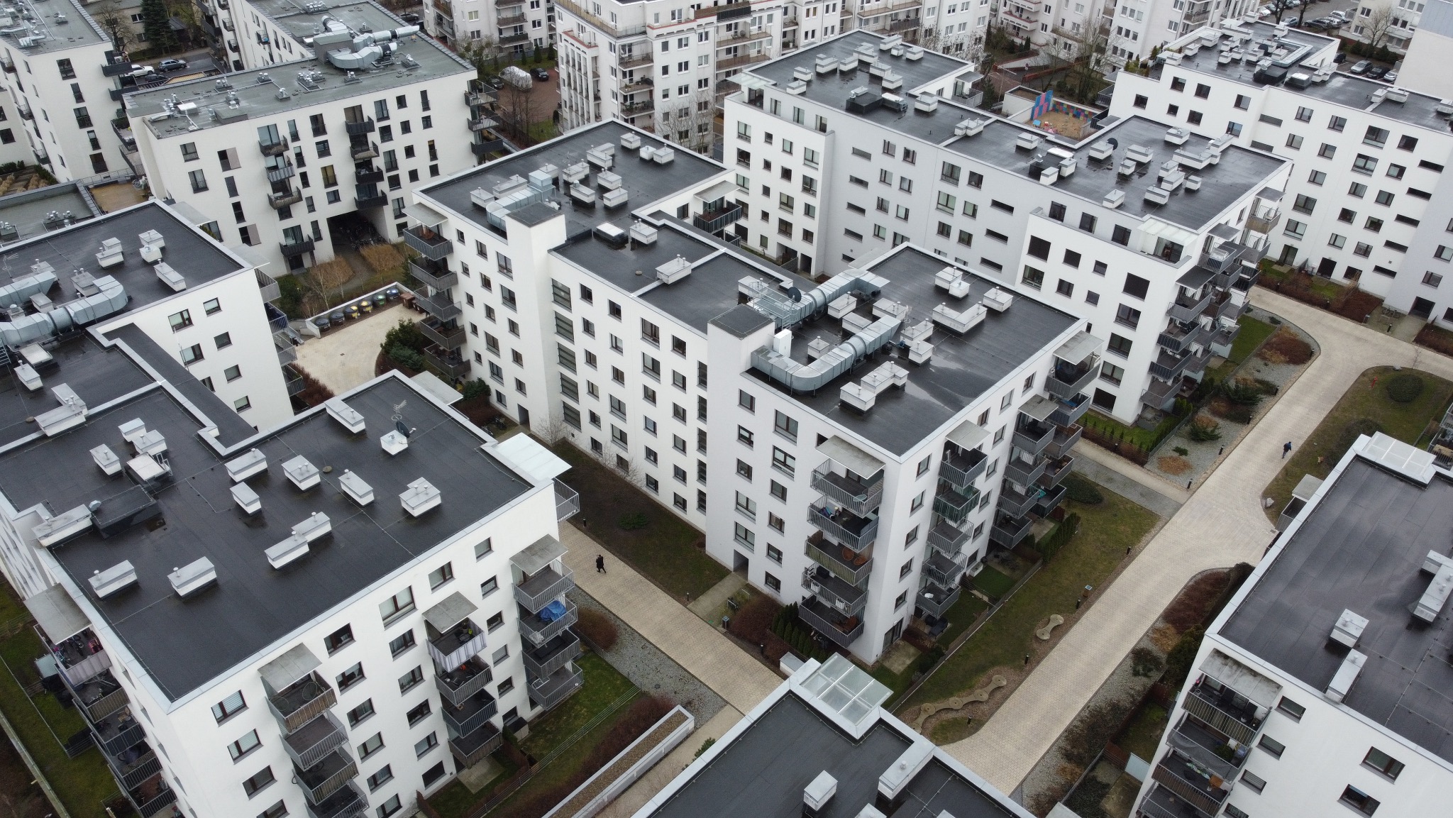 Nowoczesne, białe budynki wielorodzinne z balkonami, widok z góry. Na dachach widoczne elementy wentylacji. Pomiędzy budynkami ścieżki i zieleń.