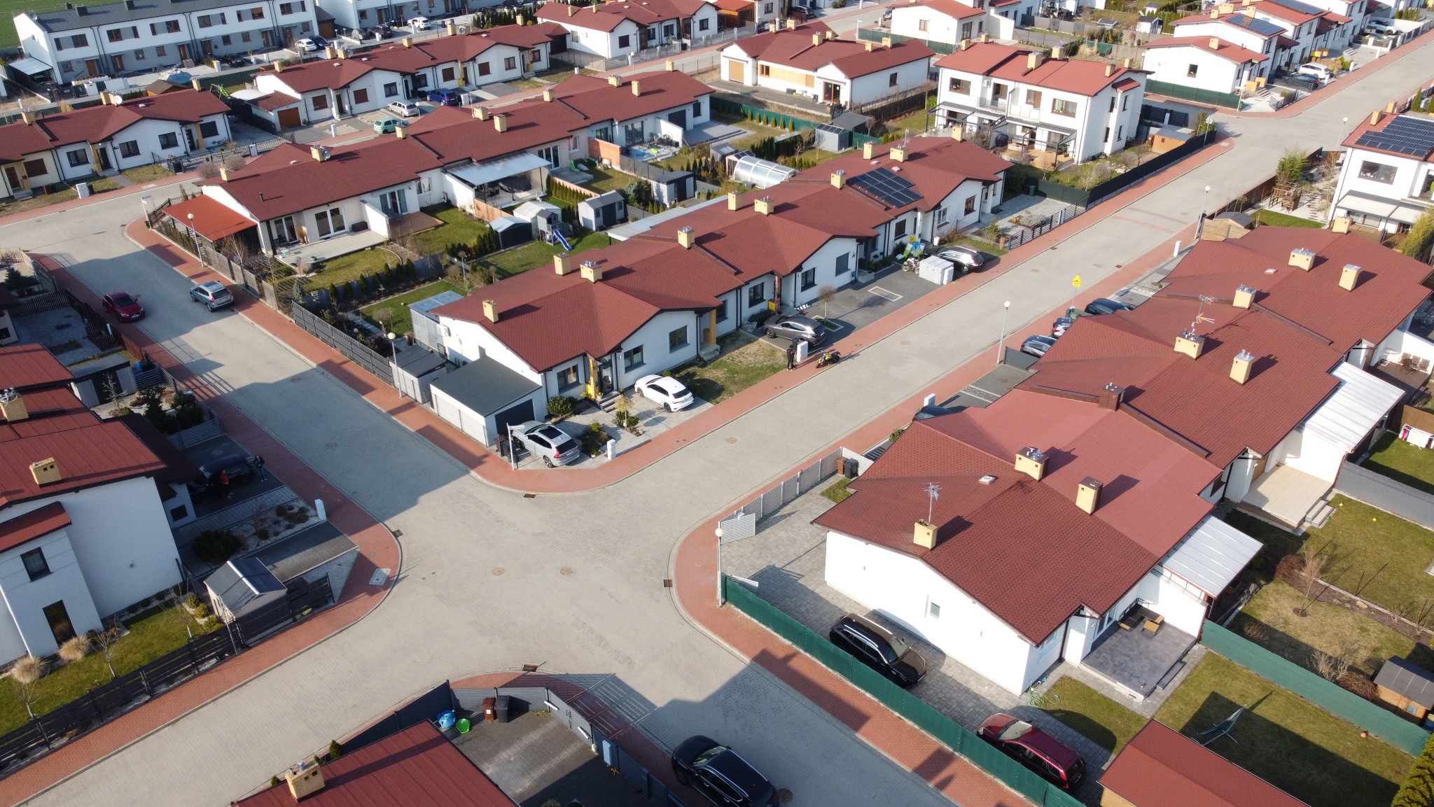 Osiedle domów jednorodzinnych z czerwonymi dachami, widok z góry. Uliczki z kostki brukowej, zadbane ogródki, samochody na podjazdach. Słoneczny dzień.