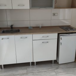 ShinEx Cleaning Service - Biała, minimalistyczna kuchnia z drewnianym blatem, płytą grzewczą i małą lodówką. Widoczne płytki na ścianie i jasna podłoga imitująca drewno.