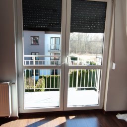 ShinEx Cleaning Service - Pokój z parkietem i oknem balkonowym z roletami. Widok na balkon i budynek mieszkalny w tle. Promienie słoneczne na podłodze.