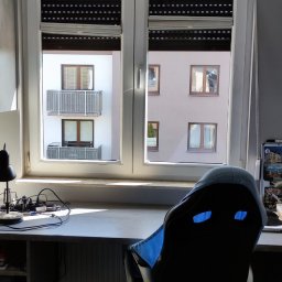 ShinEx Cleaning Service - Pokój z biurkiem i krzesłem na tle okna z roletami, przez które widać budynek z balkonami. Na biurku lampka i akcesoria komputerowe.