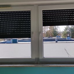 ShinEx Cleaning Service - Białe okno PCV z częściowo zasłoniętymi roletami zewnętrznymi, przez które widać budynki i drzewa. Widok z wnętrza mieszkania na zewnątrz.