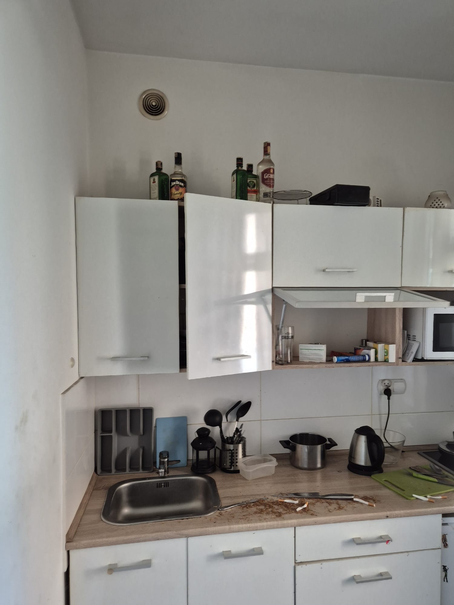 Niedomyta kuchnia z otwartymi szafkami, na blacie resztki tytoniu i niedopałki, naczynia, czajnik. Widoczne butelki alkoholu na szafkach.