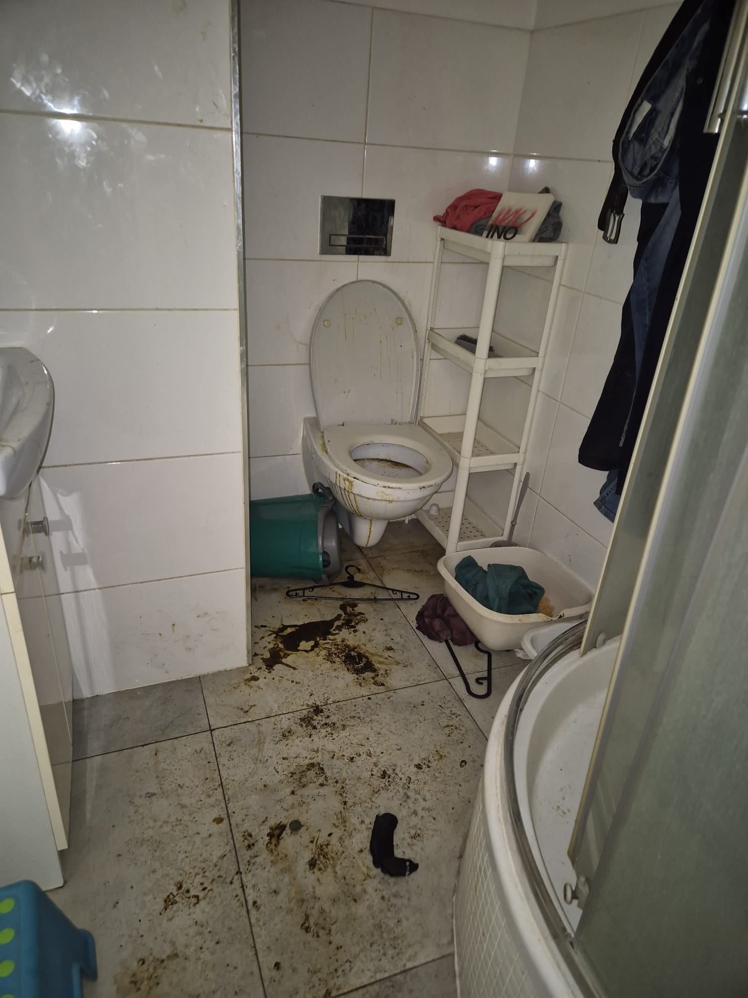 Zaniedbana łazienka z brudną toaletą, podłogą i ścianami. Widoczny przewrócony kosz, ubrania i akcesoria. Potrzeba gruntownego sprzątania.