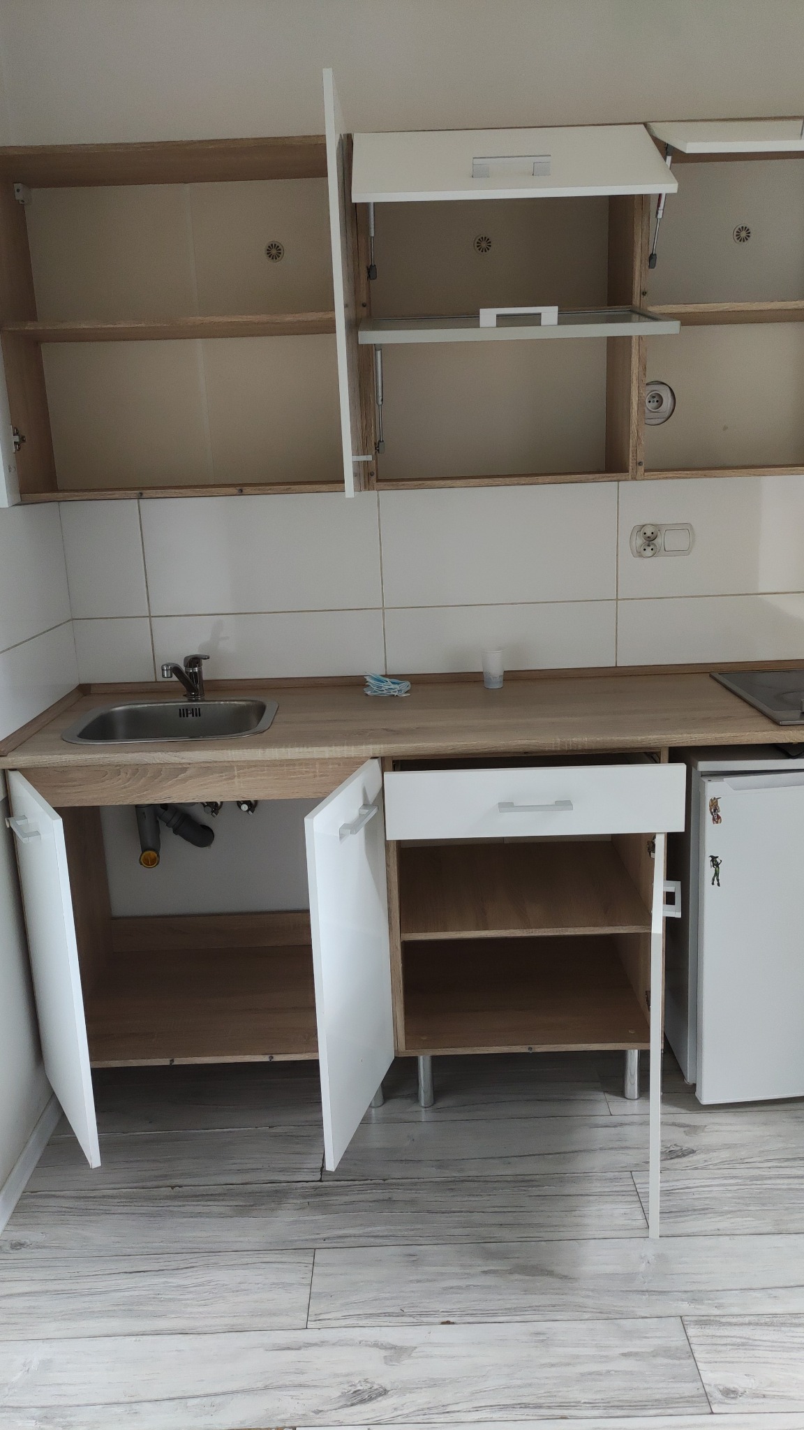 Kuchnia z otwartymi szafkami i widocznym zlewem, przygotowana do sprzątania. Płytki na ścianie i drewnopodobna podłoga. Widoczne akcesoria do mycia.