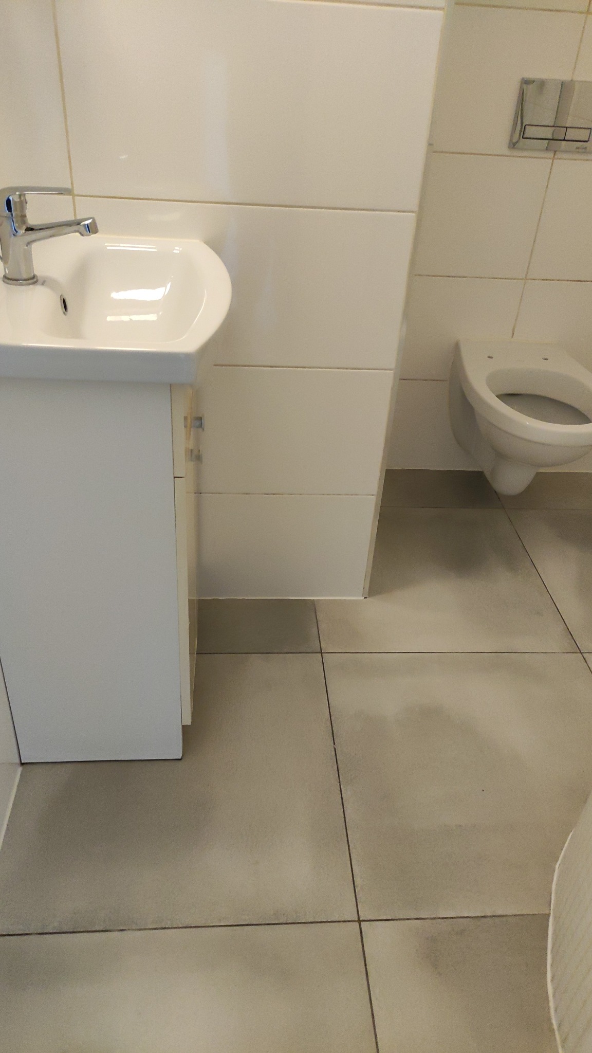 Czysta, minimalistyczna łazienka z białą umywalką, toaletą i płytkami. Widoczne ślady po umyciu podłogi, podkreślające świeżość i dbałość o detale.