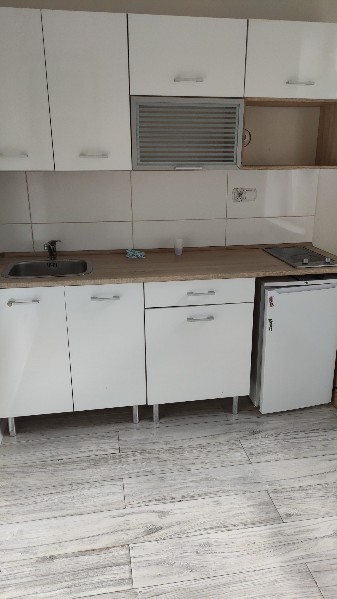 Biała, minimalistyczna kuchnia z drewnianym blatem, płytą grzewczą i małą lodówką. Widoczne płytki na ścianie i jasna podłoga imitująca drewno.