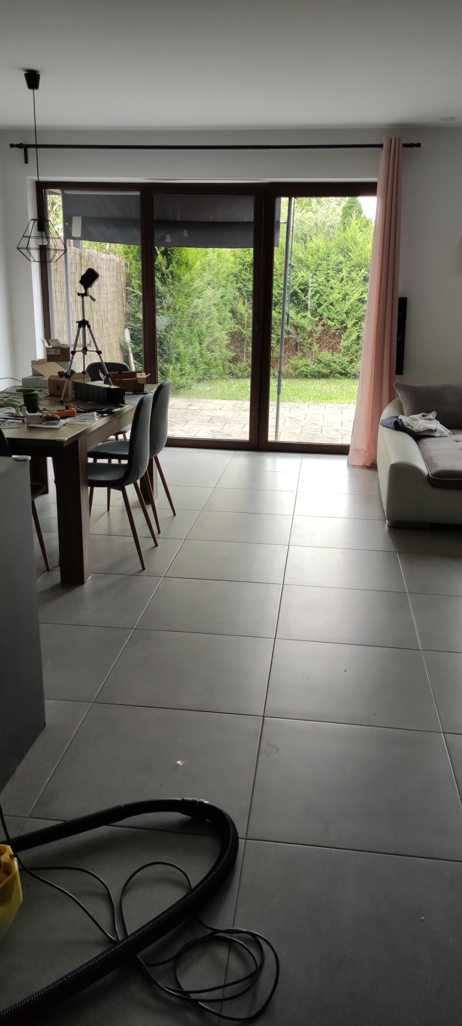 Jasne wnętrze z szarymi płytkami podłogowymi, częściowo widoczny stół z krzesłami, sofa i rura odkurzacza na podłodze. Widok na ogród przez duże okno.