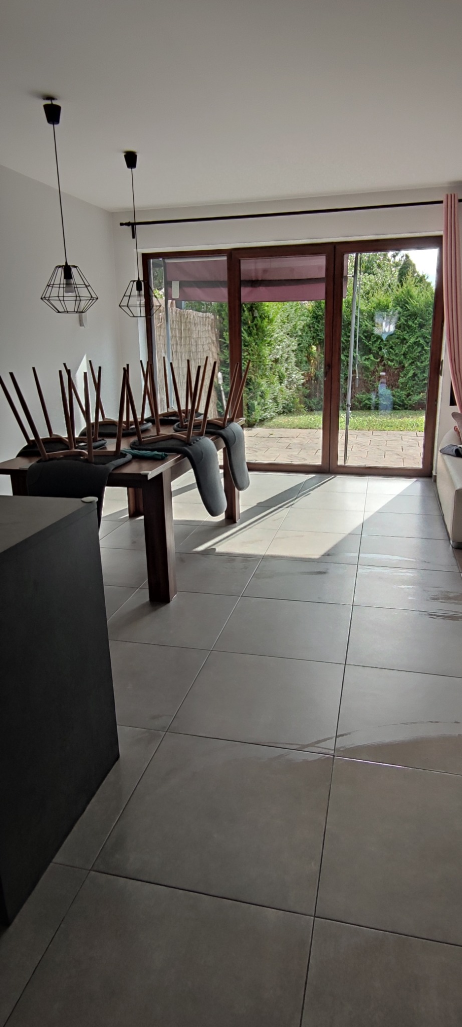 Salon z odwróconymi krzesłami na stole, przygotowany do sprzątania. Widoczne duże okno z widokiem na ogród i mokra podłoga po myciu.