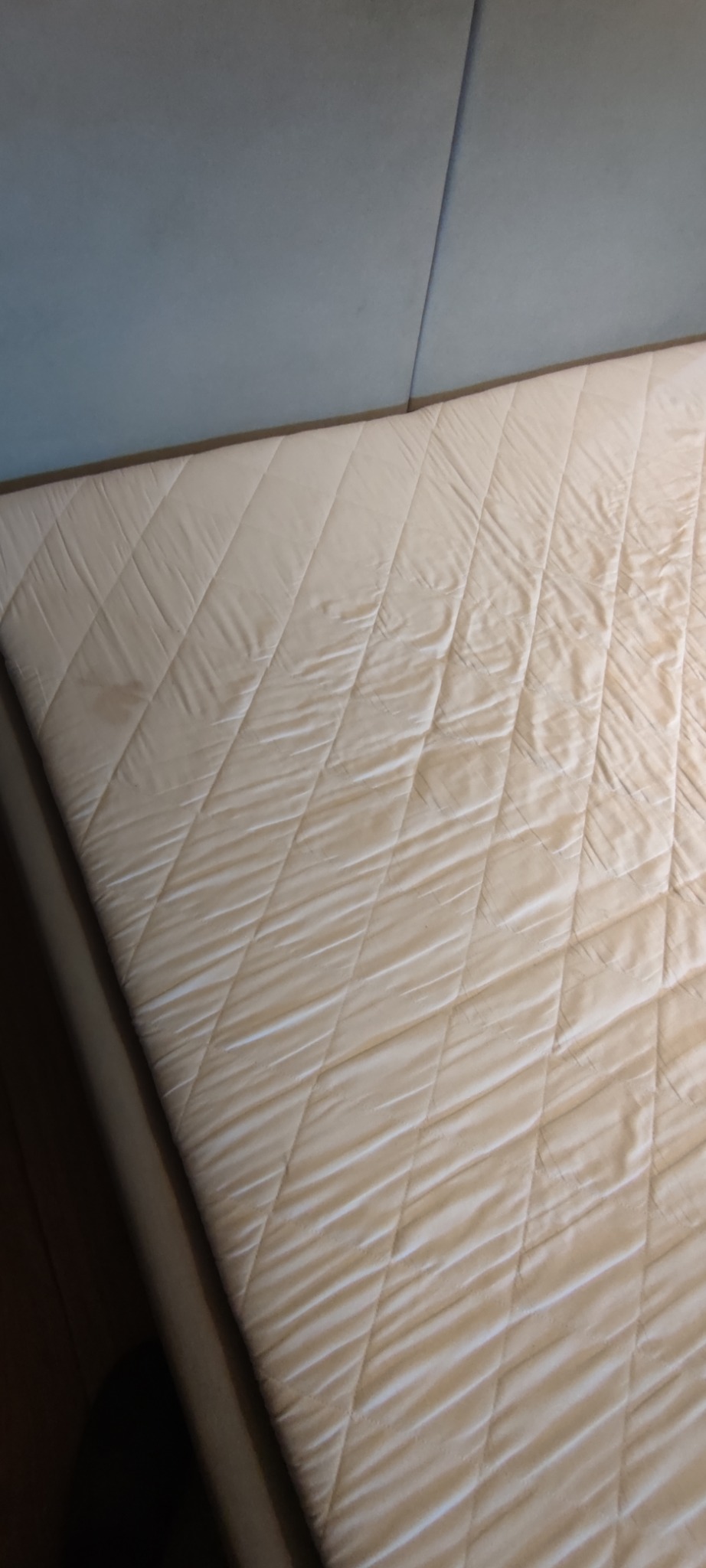 Beżowy materac z widocznymi plamami na tle niebieskiej ściany. Materac z pikowaniem w romby, ukazujący potrzebę odświeżenia i czyszczenia.