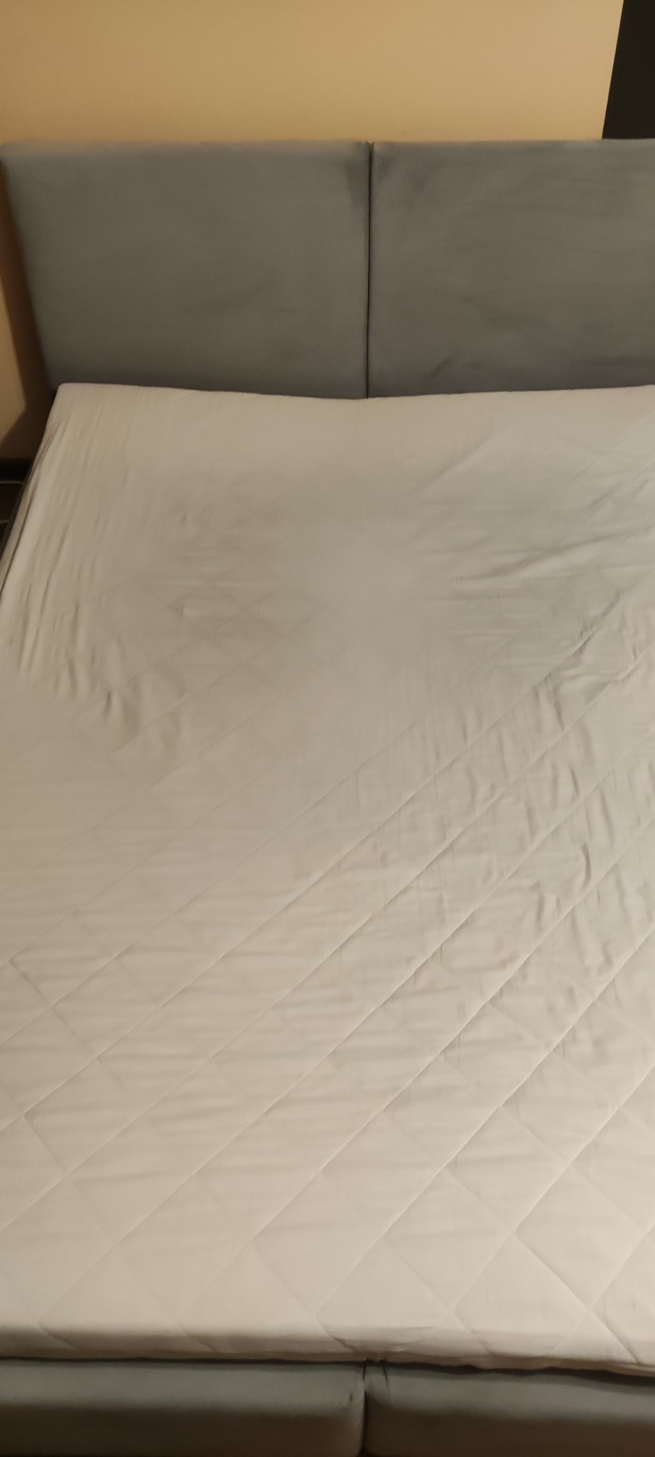 Czysty, biały materac na łóżku z szarym zagłówkiem. Widoczne pikowania w romby na materacu. Neutralne oświetlenie.