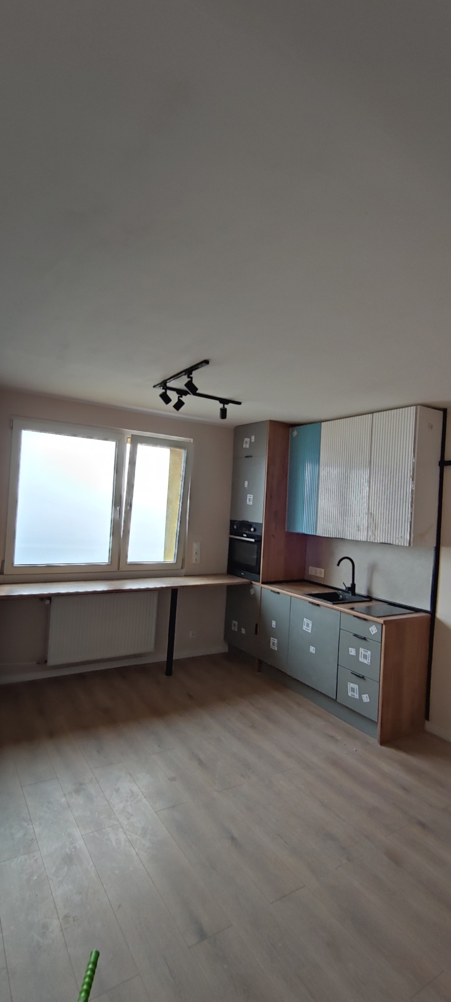 Jasna kuchnia z szarymi szafkami, drewnianym blatem i oknem z widokiem na zewnątrz. Minimalistyczny design, oświetlenie punktowe.