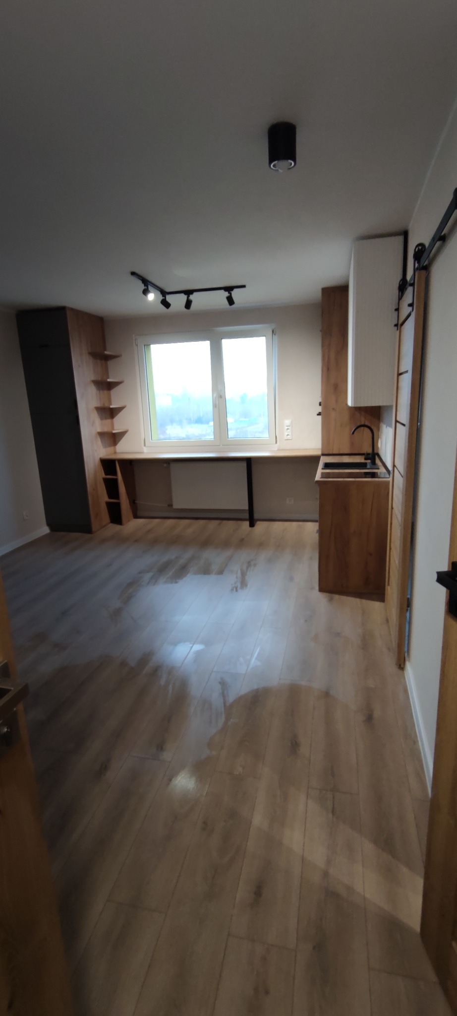 Jasne wnętrze po remoncie: drewniana podłoga, zabudowa kuchenna z czarną armaturą, biurko pod oknem, oświetlenie punktowe. Minimalistyczny design.