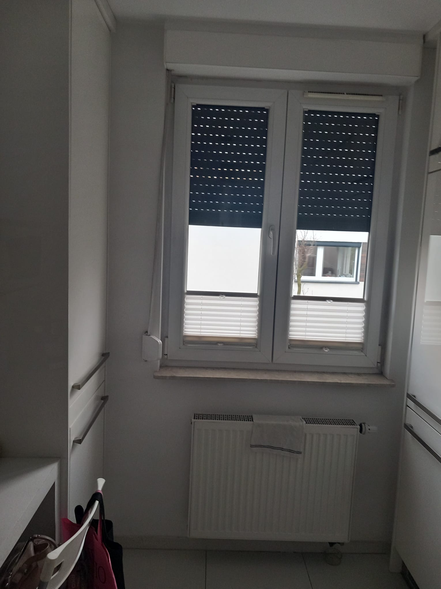 Wnętrze pokoju z oknem z roletami i plisami, biały grzejnik z ręcznikiem, szafa w zabudowie. Widok z dołu, neutralne oświetlenie.