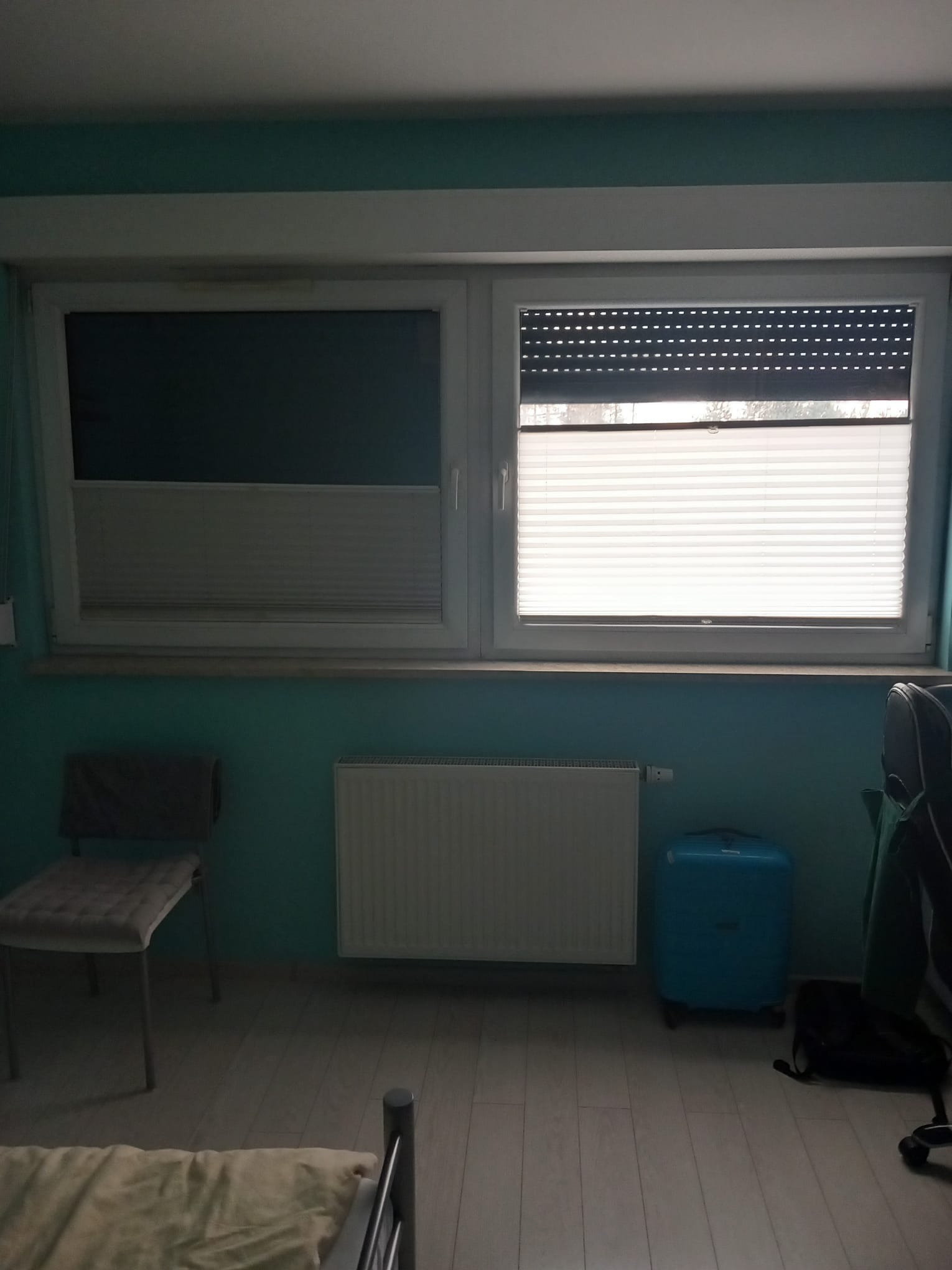 Pokój z dwoma oknami: jedno z roletą plisowaną, drugie z roletą zewnętrzną. Widoczny grzejnik, krzesło i walizka. Ściany w kolorze turkusowym.