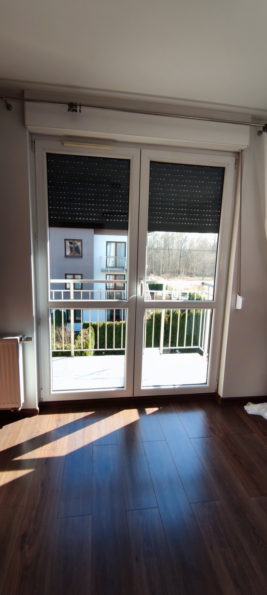 Pokój z parkietem i oknem balkonowym z roletami. Widok na balkon i budynek mieszkalny w tle. Promienie słoneczne na podłodze.